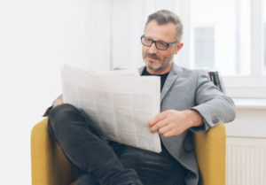 Un homme avec des lunettes se détend dans une location GuestLee lumineuse et élégante à Genève, lisant un journal dans un fauteuil luxueux près de la fenêtre. | Location de luxe GuestLee Suisse